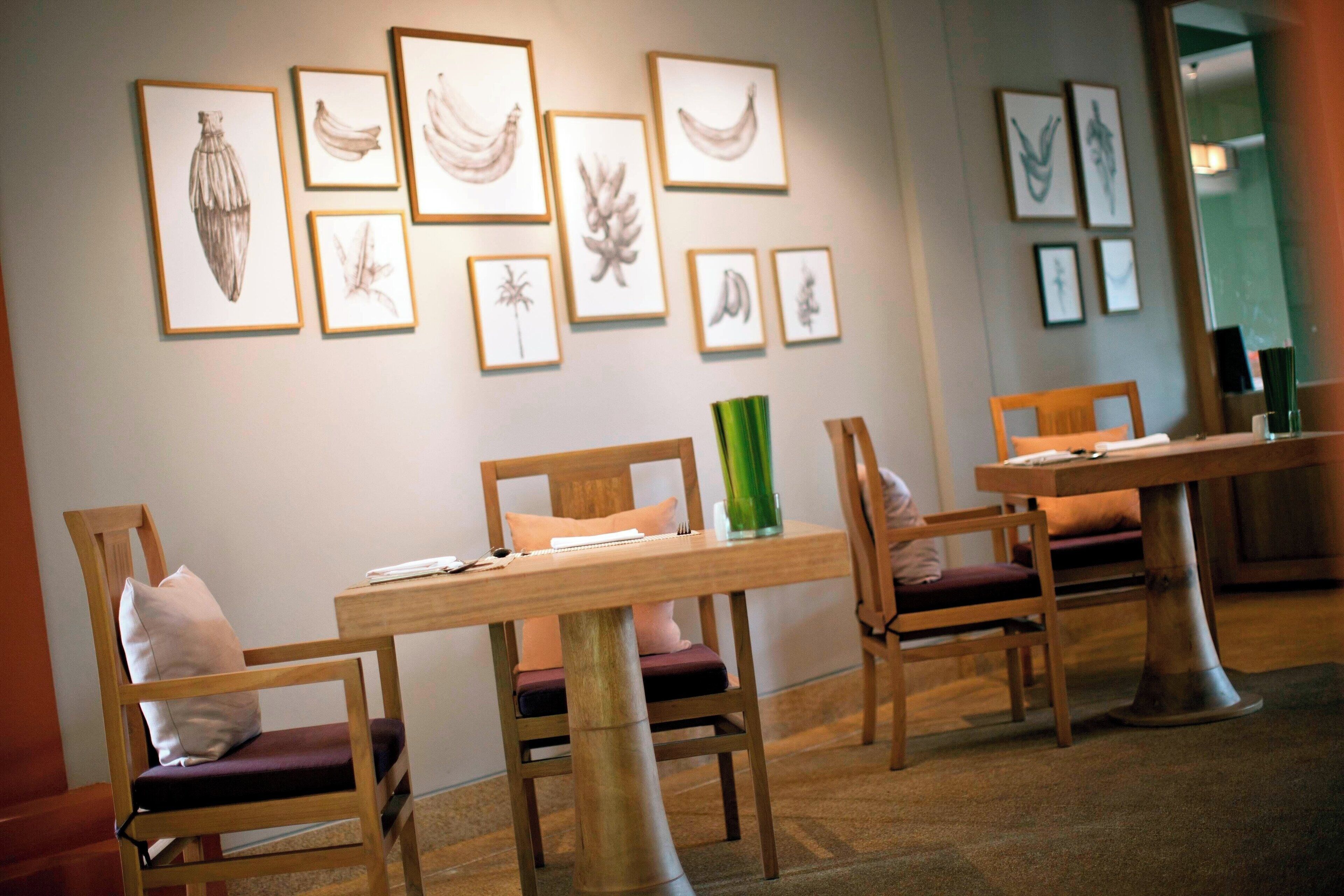 A dining area with wooden chairs and tables, framed artwork on the wall, and a green vase on one table.