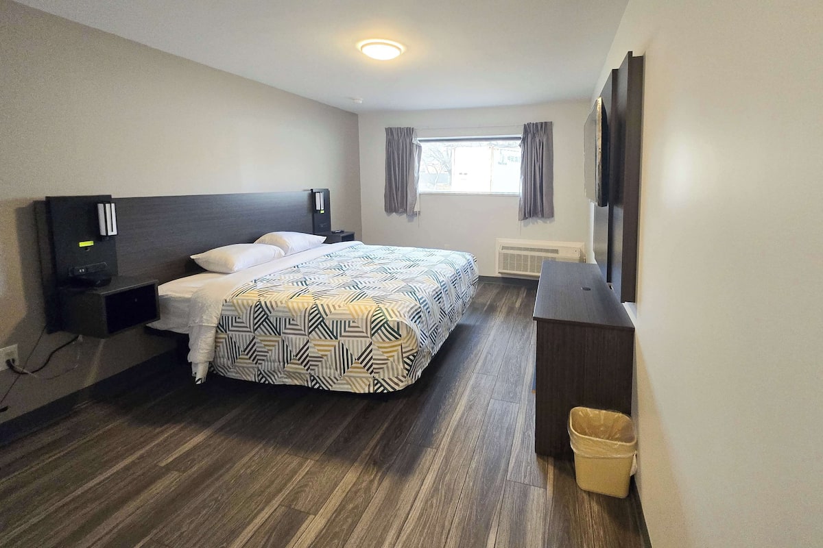 A hotel room with a bed, a desk, a TV, a window with curtains, and a waste bin.