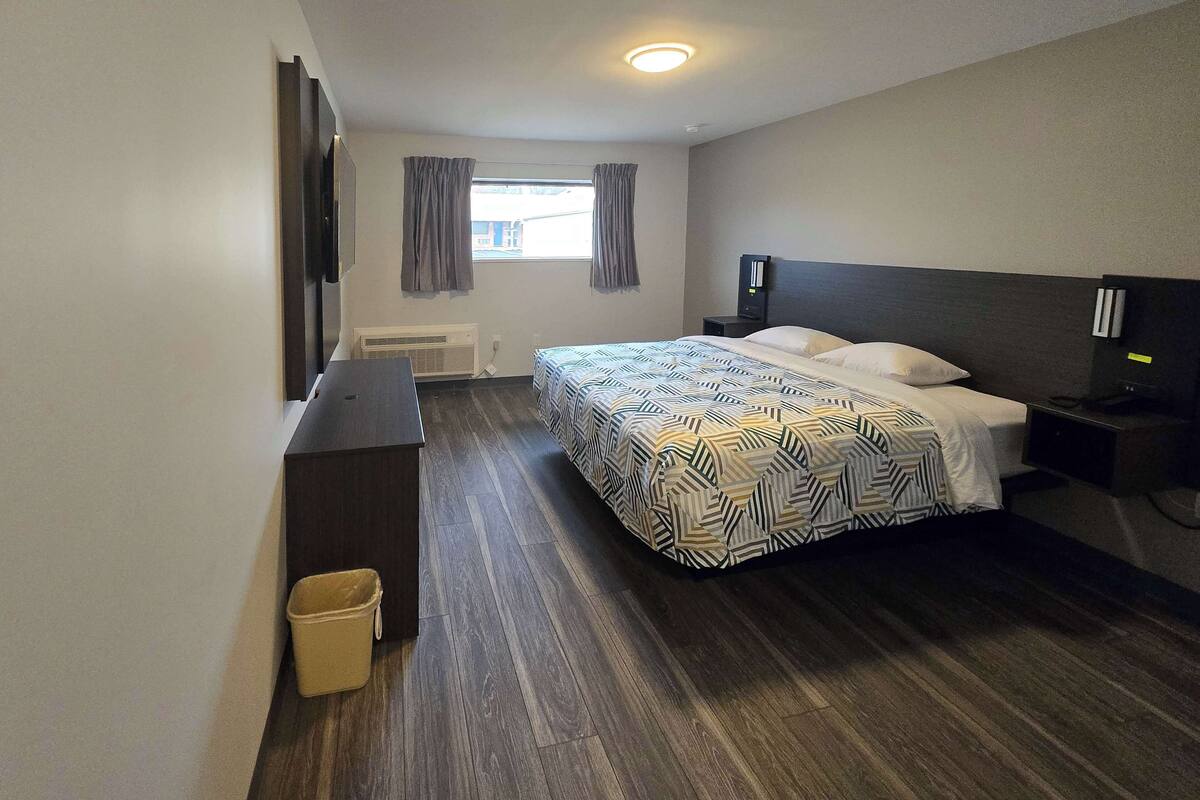 A hotel room with a bed, a desk, a chair, a waste bin, a heater, a window with curtains, and a ceiling light.