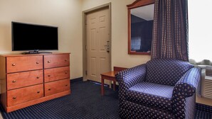 A hotel room with a wooden dresser, a television, a patterned armchair, a small table, and a door.