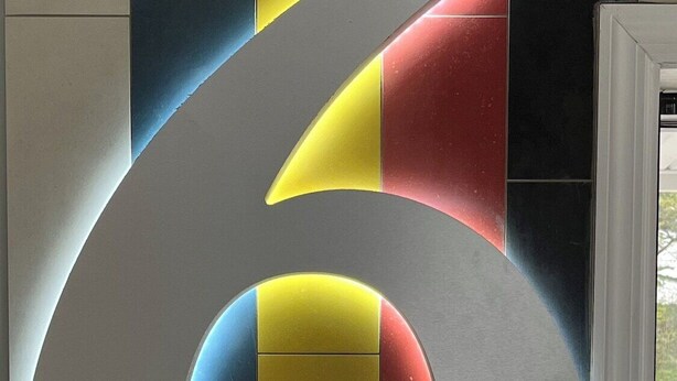 A large, metallic number 6 sculpture with a circular cutout, set against a wall with vertical stripes in blue, yellow, and red.