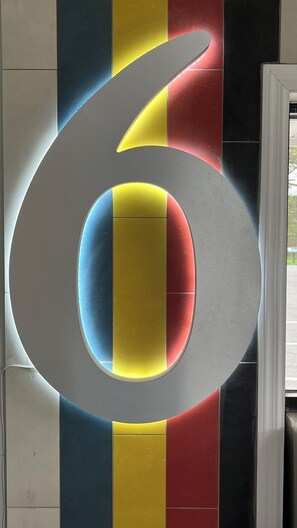 A large, metallic number 6 sculpture with a circular cutout, set against a wall with vertical stripes in blue, yellow, and red.