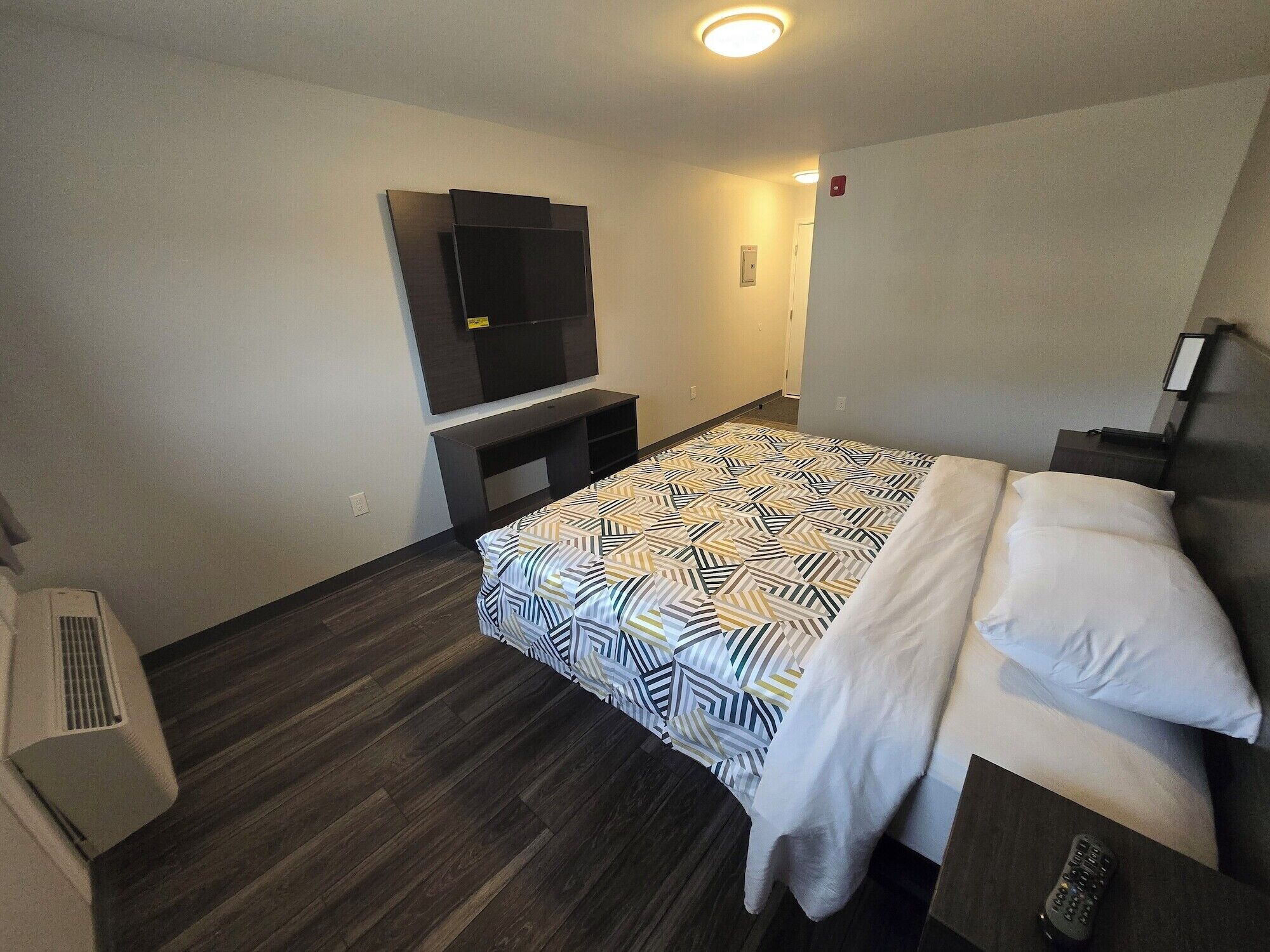 A hotel room with a bed, a TV mounted on the wall, a small table, and a wall-mounted heater.