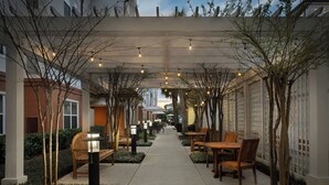 A covered outdoor seating area with tables and chairs, surrounded by trees and plants, and a pathway leading to a building entrance. - Residence Inn by Marriott Sandestin at Grand Boulevard (Miramar Beach)