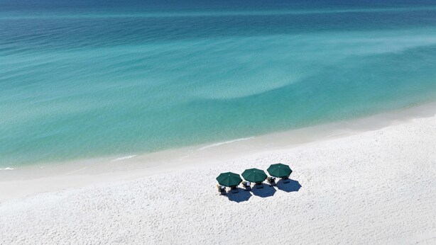 A beach with white sand and a clear blue sea. There are three green umbrellas and chairs on the sand. - Residence Inn by Marriott Sandestin at Grand Boulevard (Miramar Beach)