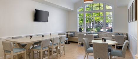 A dining area with a long table and several chairs, a large window with a view of greenery, and a flat-screen TV mounted on the wall.