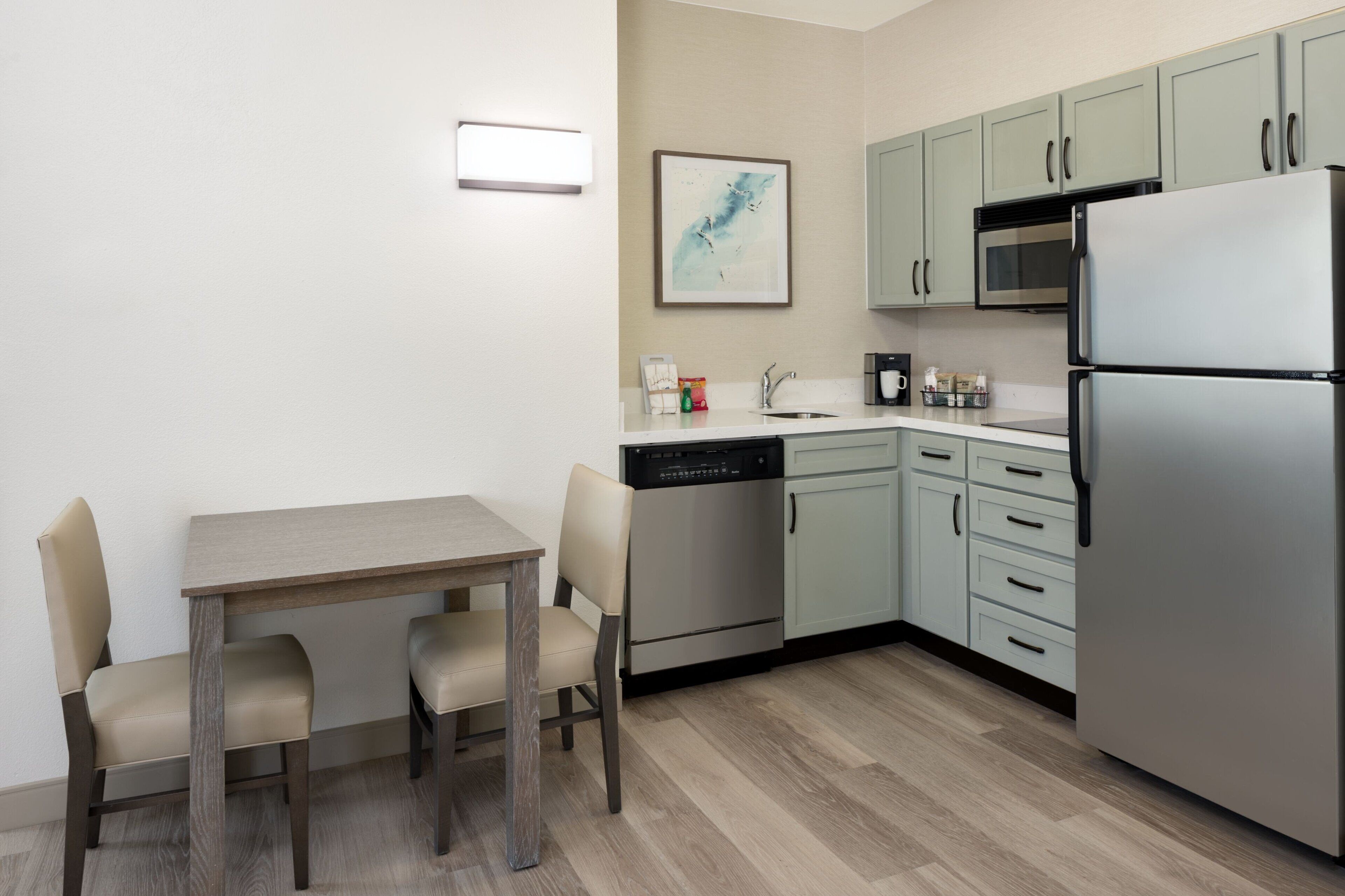 Una cocina moderna con armarios verde claro, un refrigerador de acero inoxidable, un microondas y una cafetera. Hay una mesa de comedor con dos sillas y un cuadro en la pared.