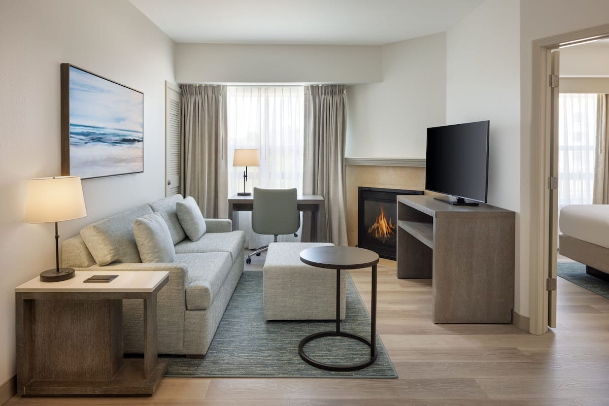 Une chambre d’hôtel moderne avec un canapé, une petite table ronde, un téléviseur, une cheminée et une vue sur l’océan par la fenêtre.