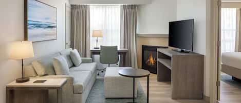 A modern hotel room with a sofa, a small round table, a TV, a fireplace, and a view of the ocean through the window.