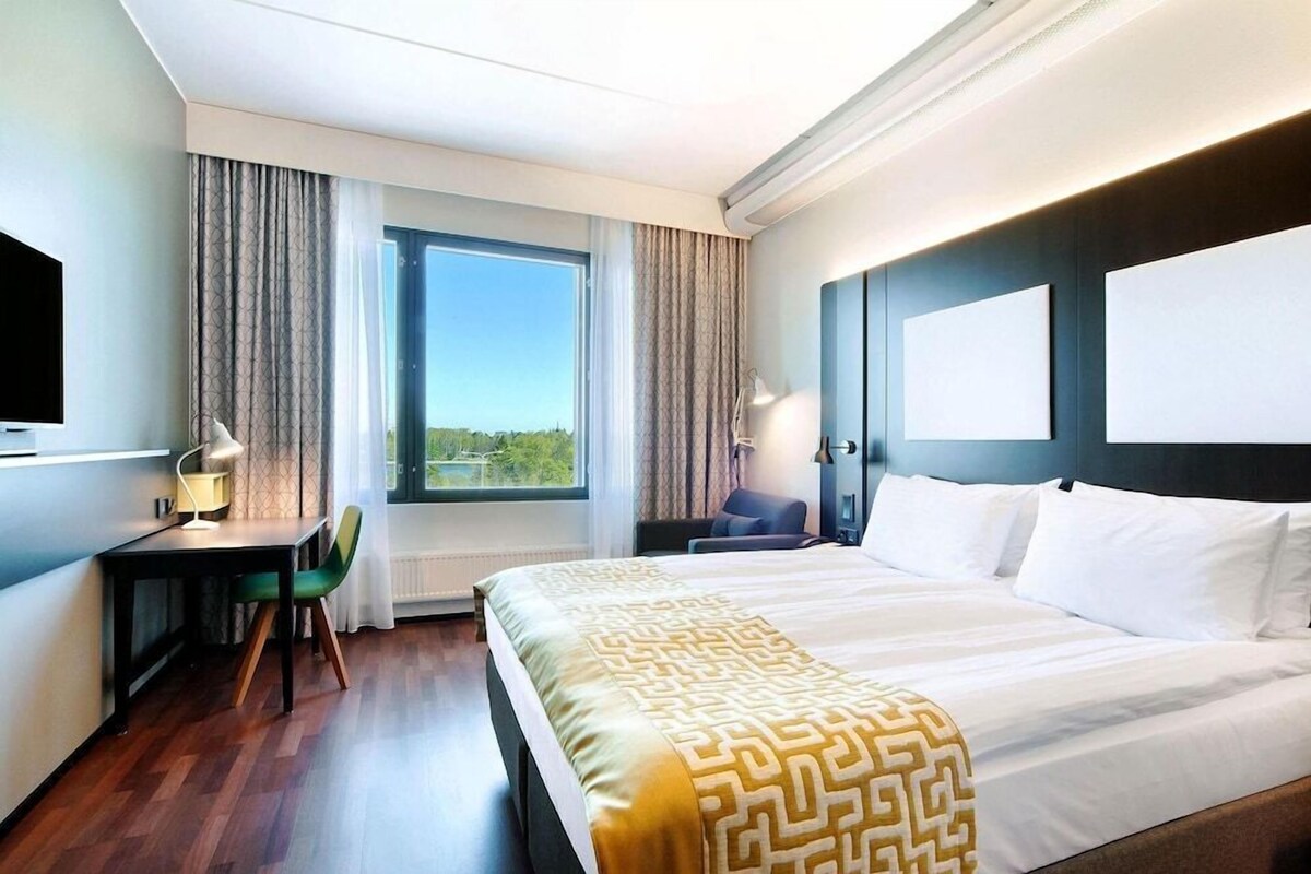 A hotel room with a bed, a desk, a chair, a TV, and a window with curtains.