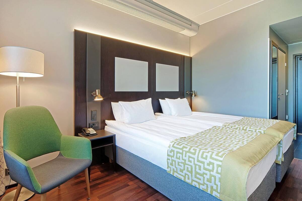 A hotel room with a bed, a chair, a bedside table with a lamp, and a door.