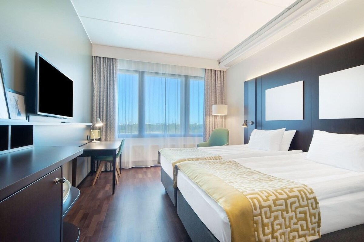 A hotel room with a bed, a desk, a chair, a TV, and a window with curtains.