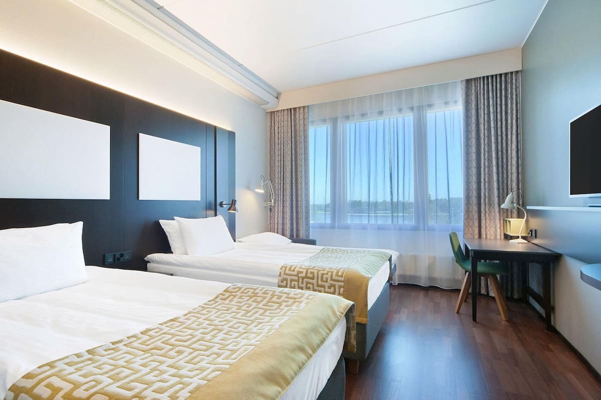 A hotel room with two beds, a desk, a chair, a large window with curtains, and a flat-screen TV.
