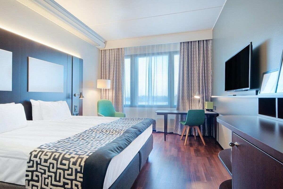 A hotel room with a large bed, a desk, two chairs, a television, and a window with curtains.