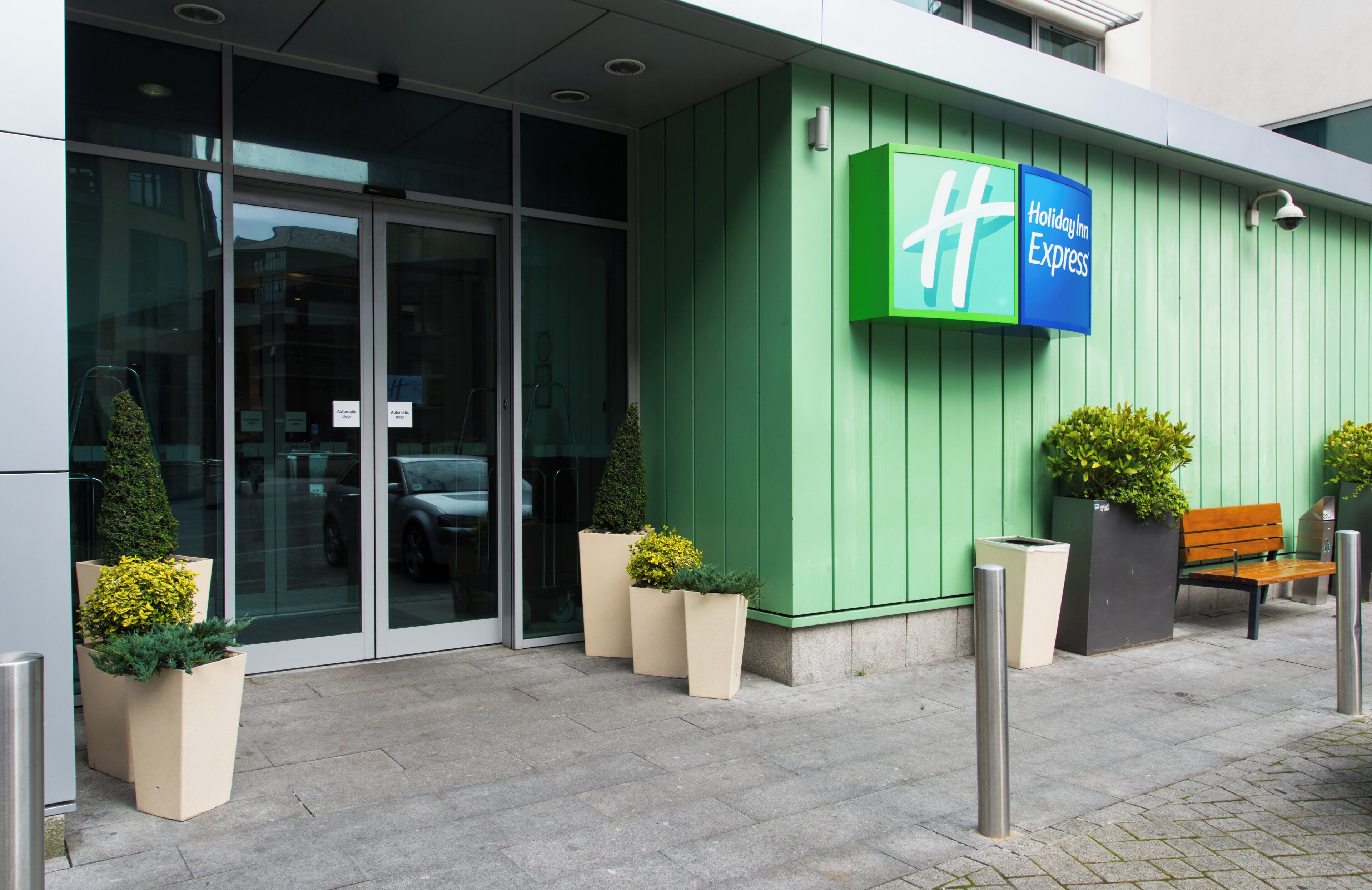 A hotel entrance with a green facade, automatic doors, and a bench outside.