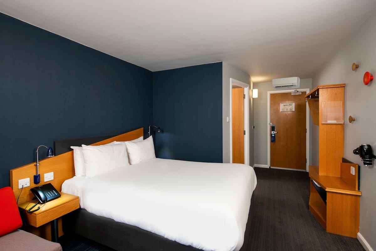 A hotel room with a large bed, a bedside table with a phone, a red chair, a wooden desk, and a wardrobe.