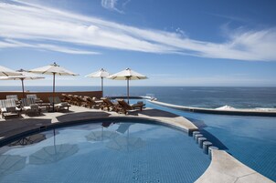 Zona junto a la piscina con sillones de descanso y sombrillas, con vistas al océano.
