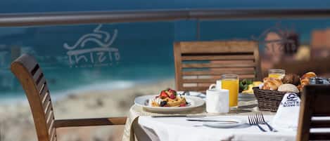 A table set with breakfast items, including waffles, fruit, and juice, overlooking a beach with a clear view of the ocean.