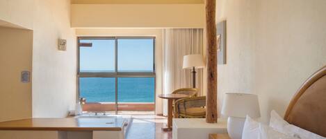A hotel room with a bed, a desk, a chair, a lamp, and a view of the sea.