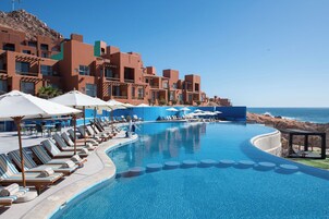 Piscina de resort con sillones de descanso y sombrillas, cielo azul despejado y vista al océano.