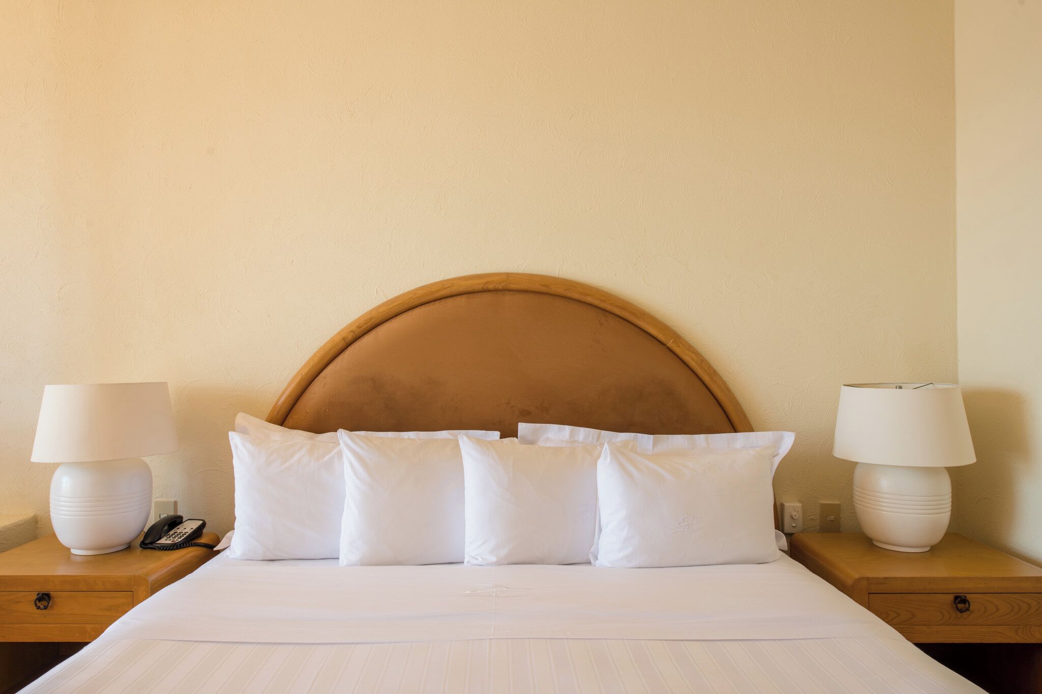 Quarto de hotel com uma cama grande, roupa de cama branca, cabeceira de madeira, duas mesas de cabeceira com abajures e um telefone.