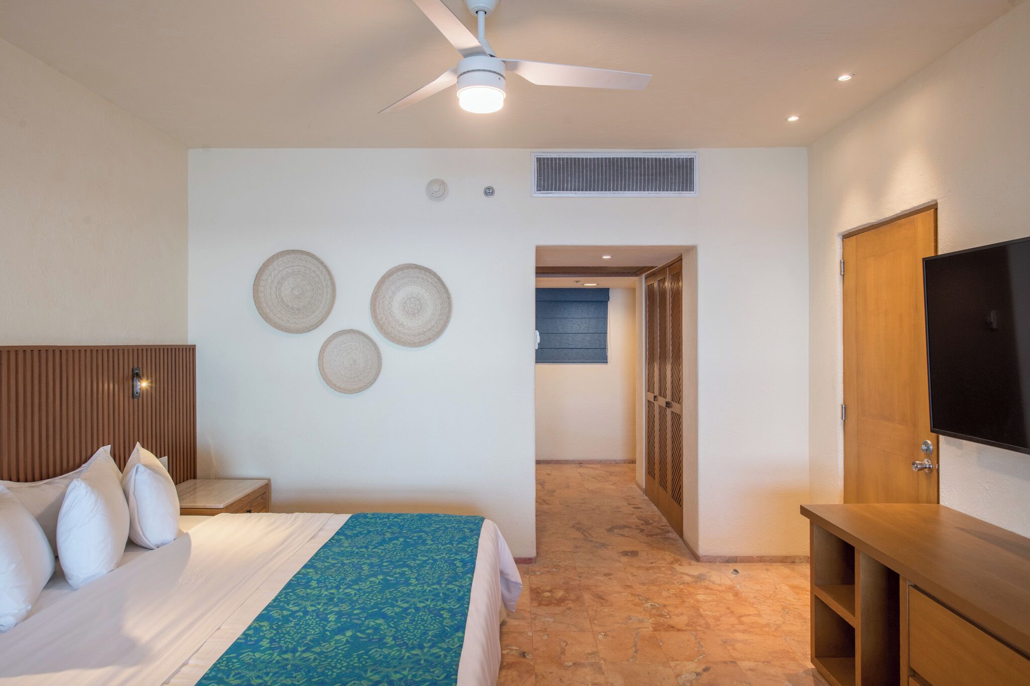 Een hotelkamer met een bed, een televisie, een plafondventilator en een gang naar een andere kamer.