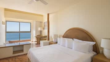 A hotel room with a large bed, a desk with a chair, a window with a view of the ocean, and a ceiling fan.
