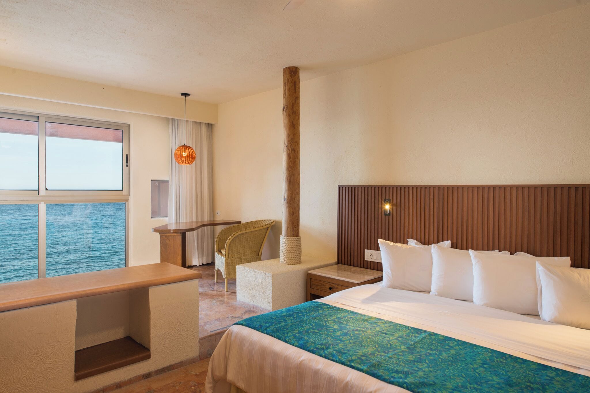 A hotel room with a large bed, a desk, a chair, a window with a view of the sea, and a wooden headboard.