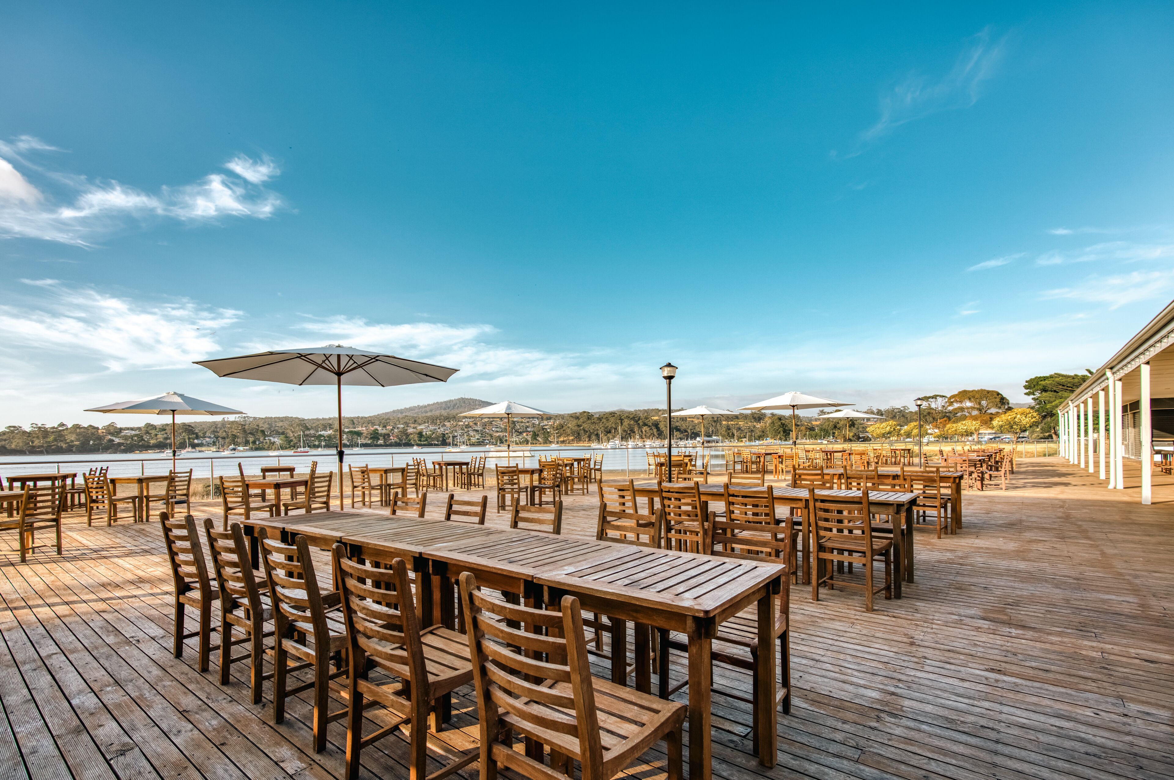 Un espacio de comedor al aire libre con mesas y sillas de madera, sombrillas y vistas a un lago.
