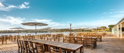Un espacio de comedor al aire libre con mesas y sillas de madera, sombrillas y vistas a un lago.