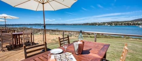 Un espace extérieur aménagé pour les repas, avec des tables et des chaises en bois, un grand parasol et une vue panoramique sur l’eau et les collines en arrière-plan.