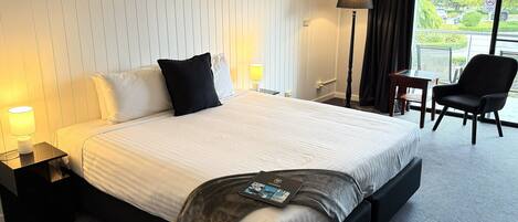Ett hotellrum med en stor säng, ett skrivbord, en stol, en lampa och utsikt över omgivningarna.