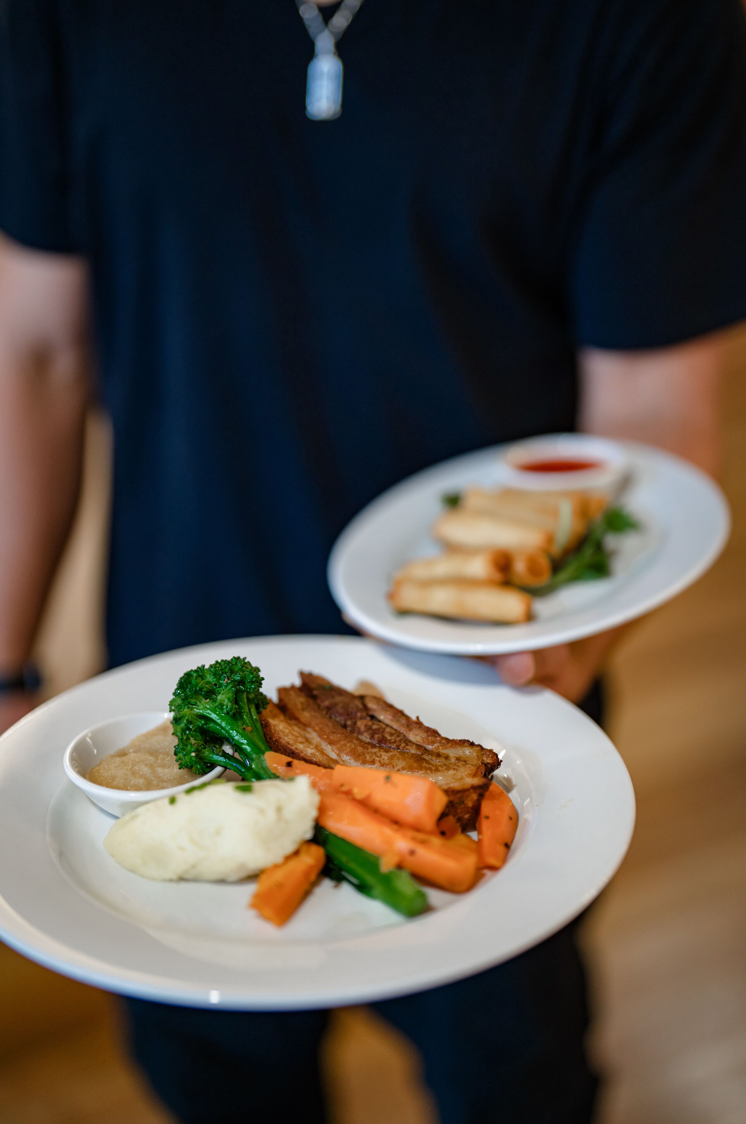 A person holding two plates of food, one with a meat dish and vegetables, and the other with spring rolls and a dipping sauce.