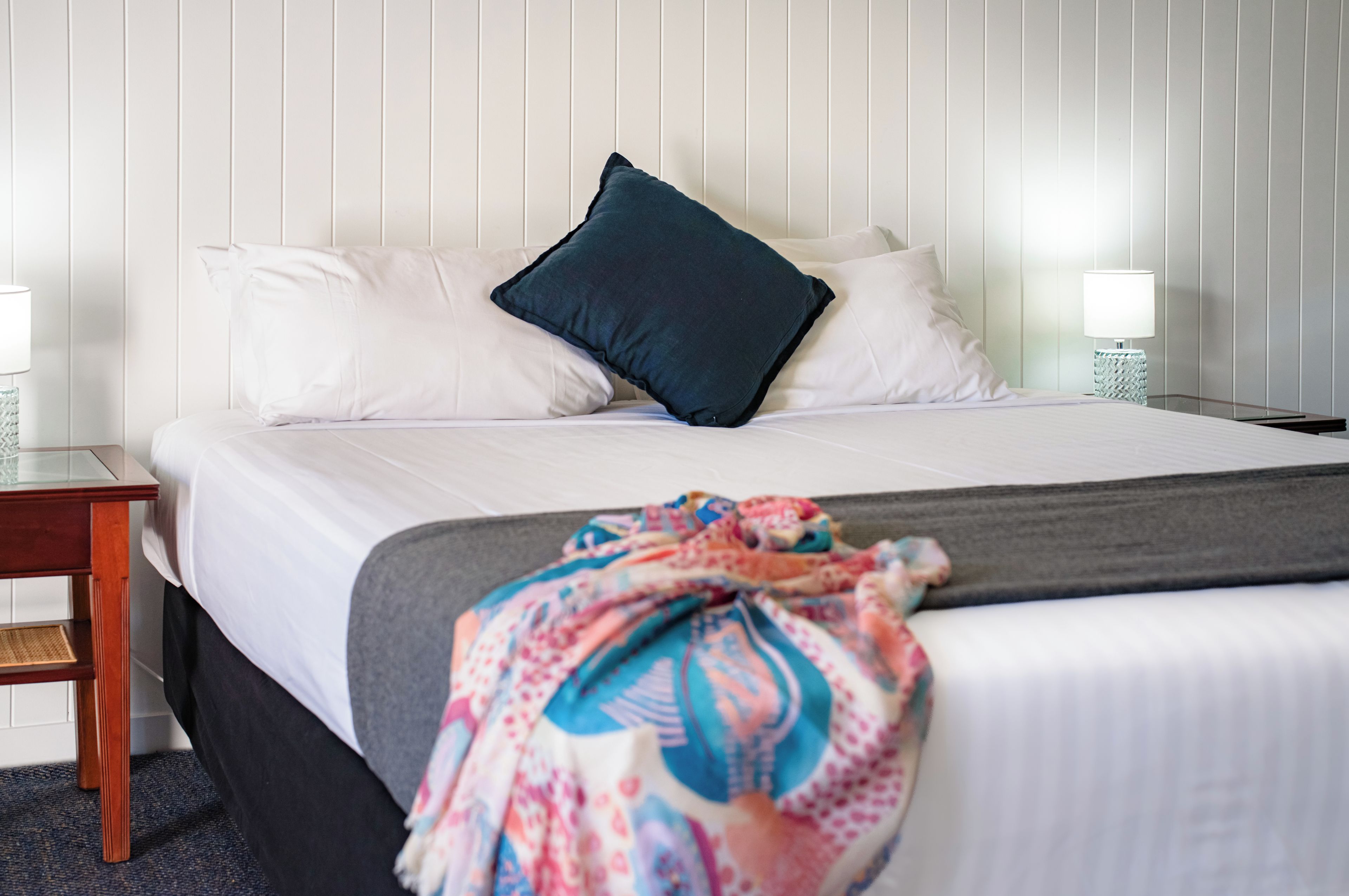 A neatly made bed with white linens and a dark blue pillow, a colorful blanket, and a bedside table with a lamp.
