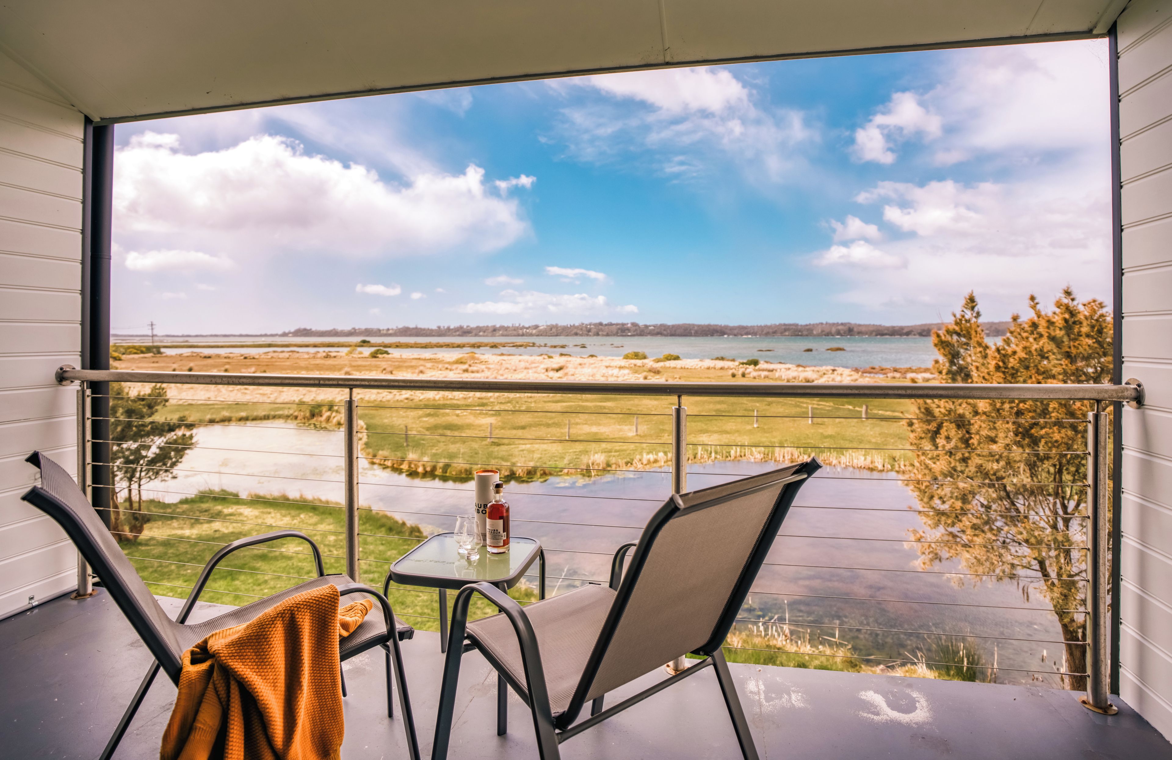 Een balkon met een tafel en stoelen, een fles en een glas, met uitzicht op een waterpartij en een verafgelegen landschap.