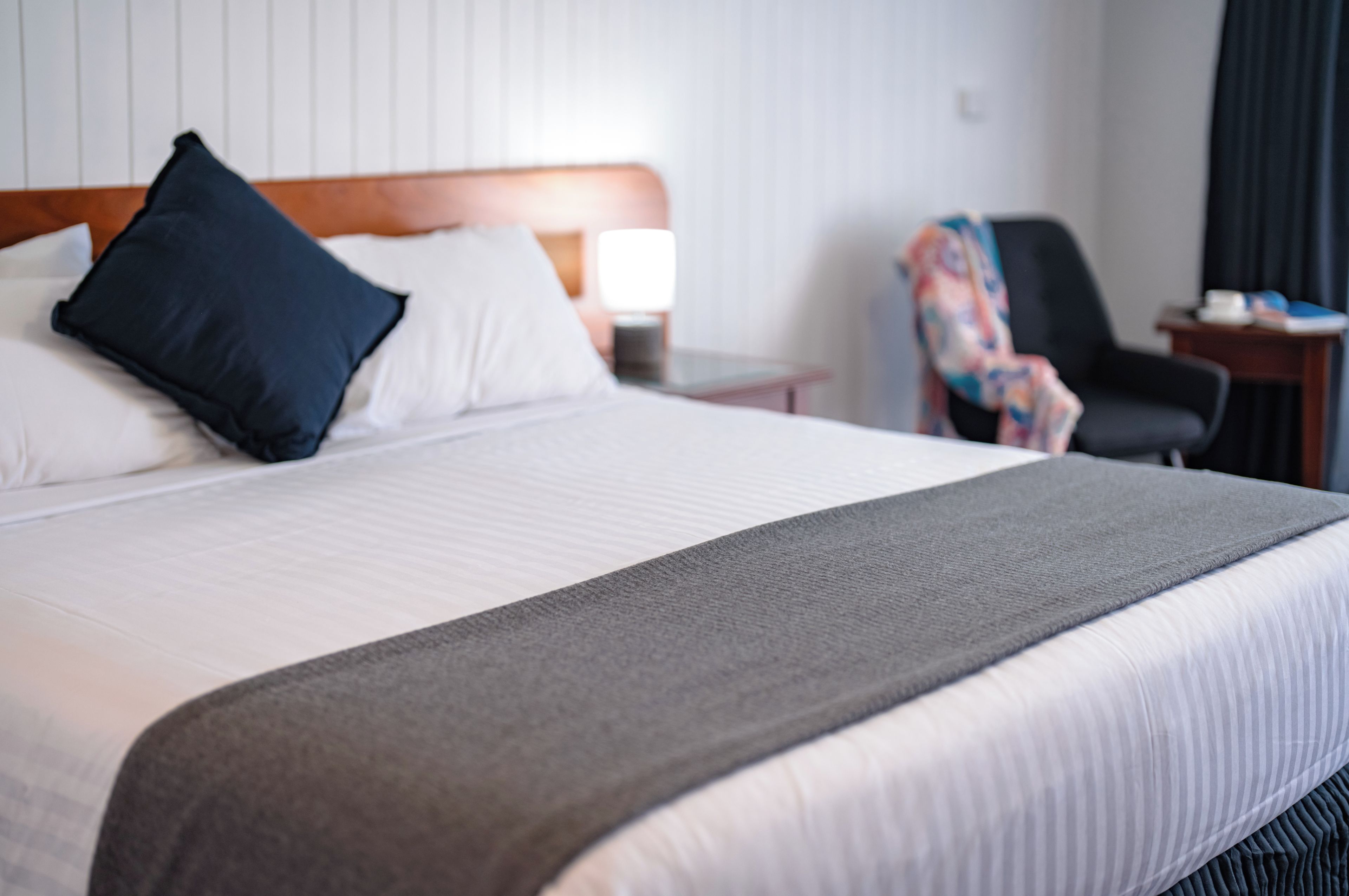 A hotel room with a bed, pillows, a nightstand, a lamp, a chair, and a table with items on it.