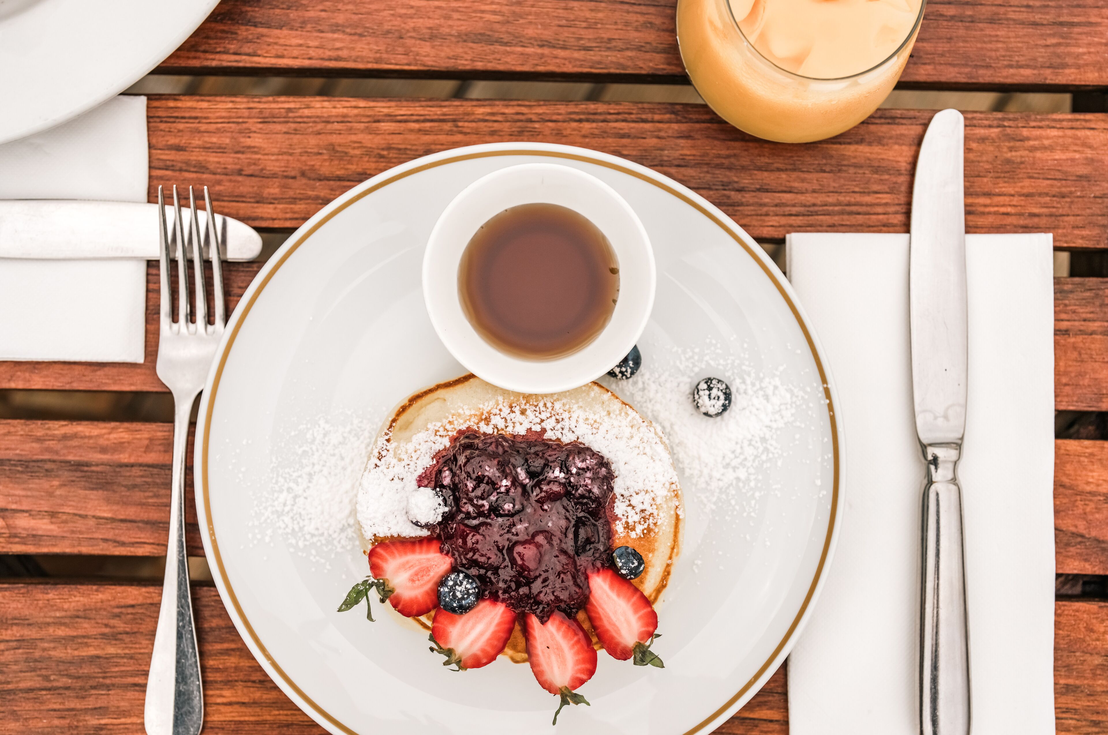 Un desayuno con tortitas cubiertas de frutos rojos y azúcar glas, una taza de café y un vaso de zumo de naranja sobre una mesa de madera.