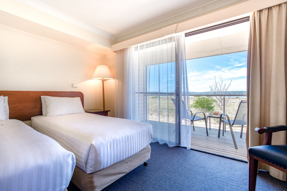 A hotel room with two beds, a nightstand with a lamp, a view of the outdoors through a large window, and a chair by the window.