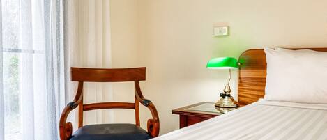 A hotel room with a bed, a wooden chair, a nightstand with a lamp, and a window with sheer curtains.