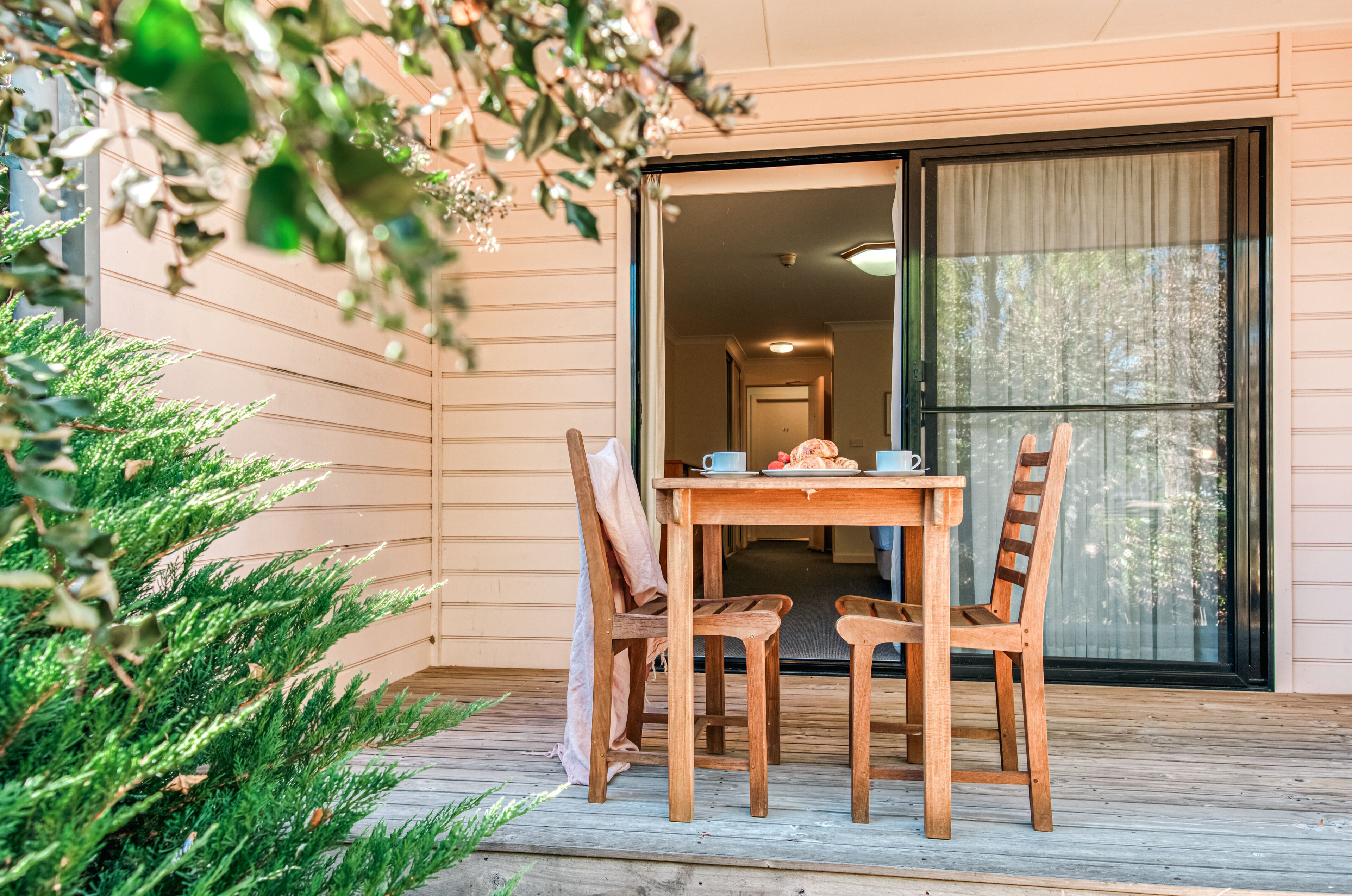 An outdoor wooden deck with a table and chairs, a potted plant, and a sliding glass door leading to a room.