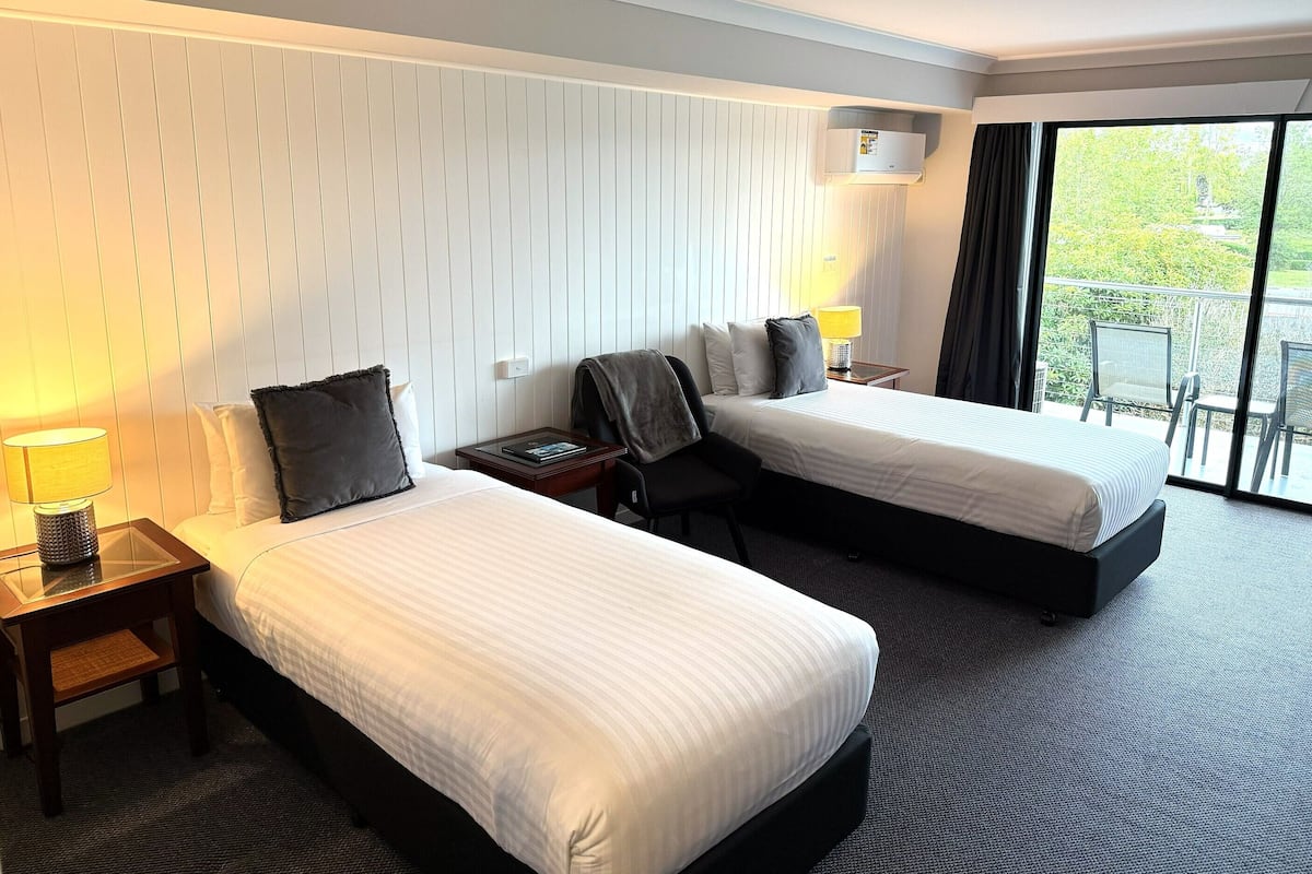 Quarto de hotel com duas camas, uma mesa de cabeceira com abajur, uma cadeira e janela com vista para o verde.