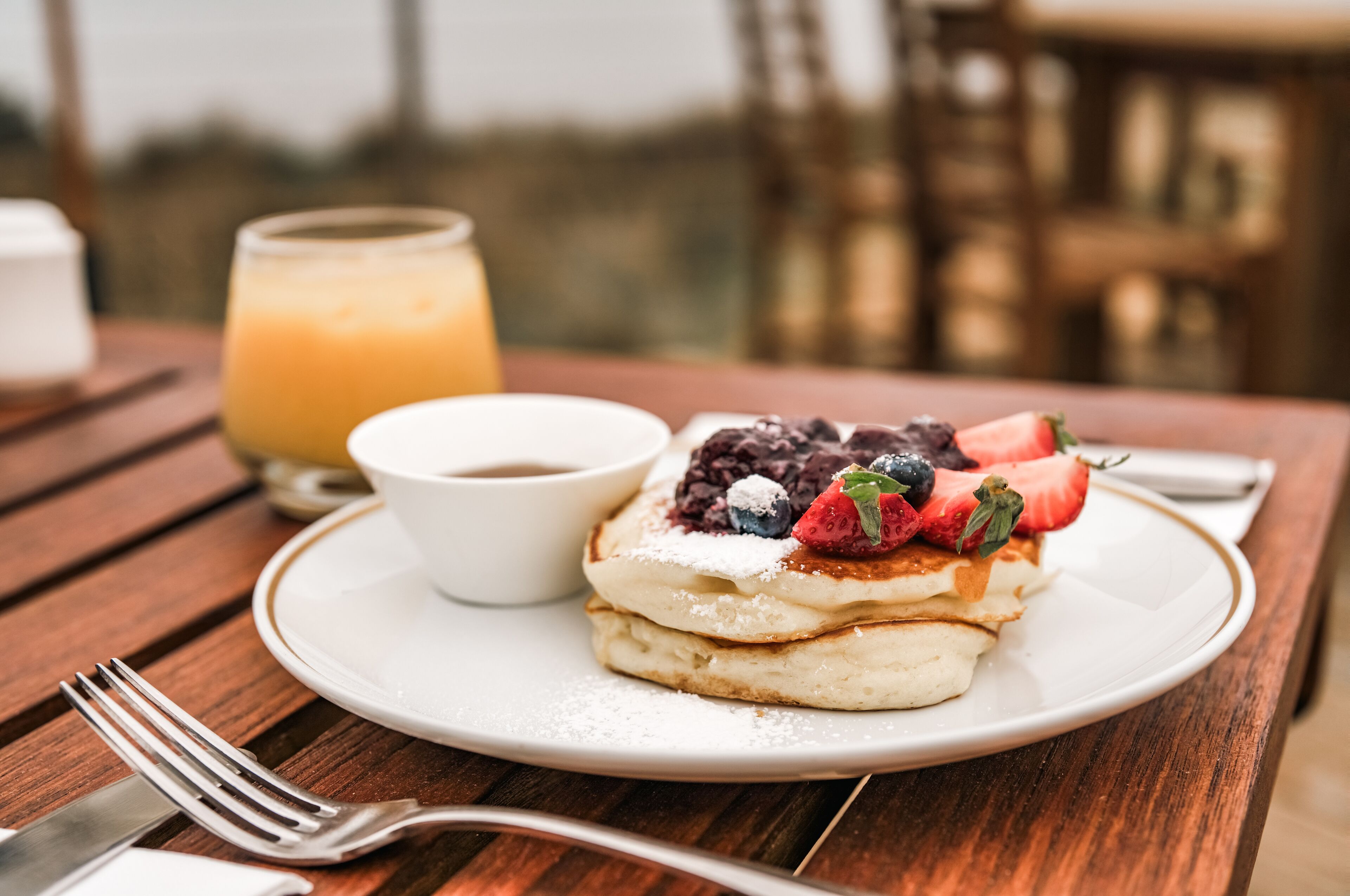 A breakfast setting with pancakes topped with berries and powdered sugar, a glass of orange juice, and a cup of coffee on a wooden table.