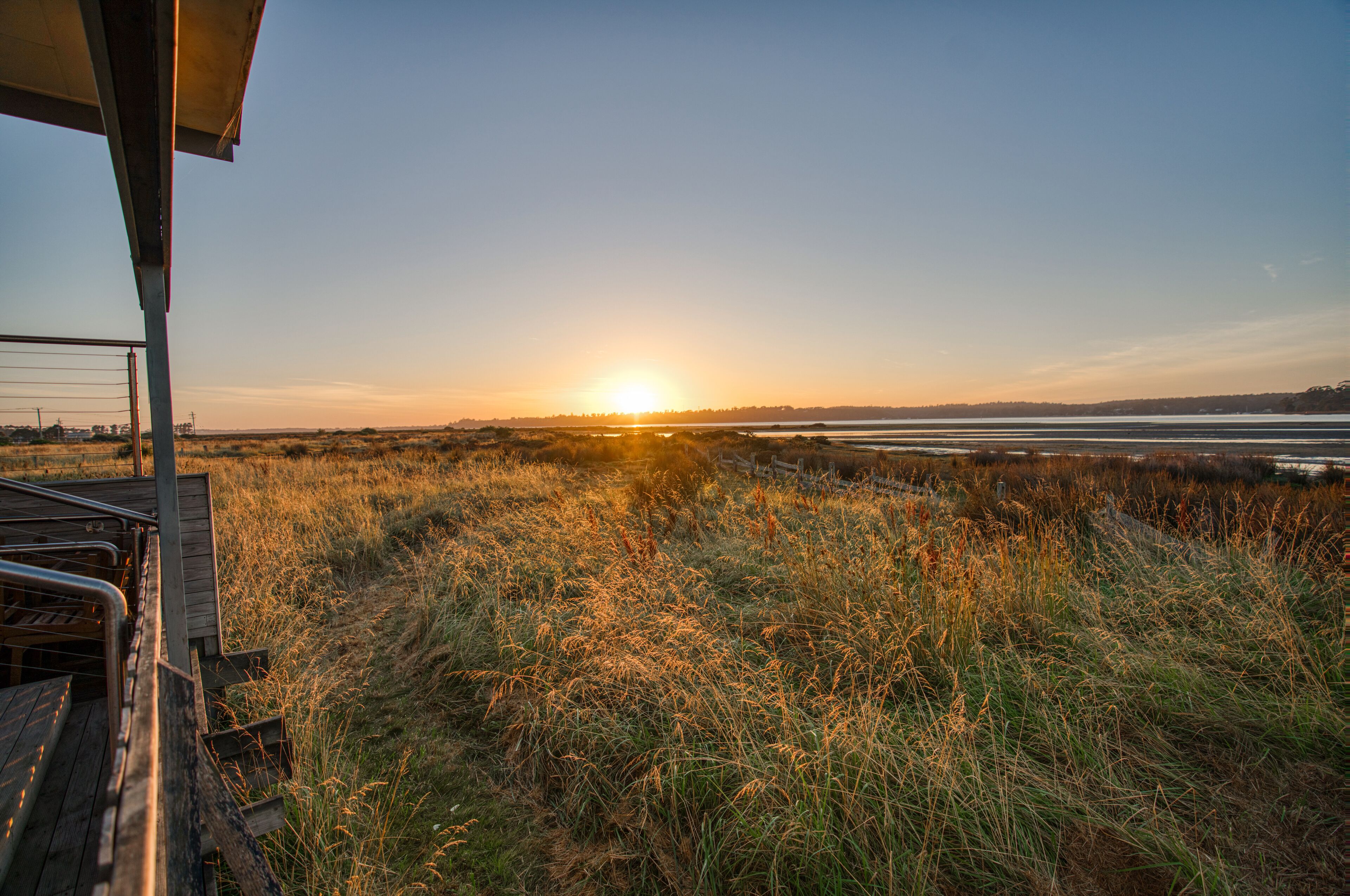 A sunset view from a wooden deck overlooking a grassy field and a body of water.