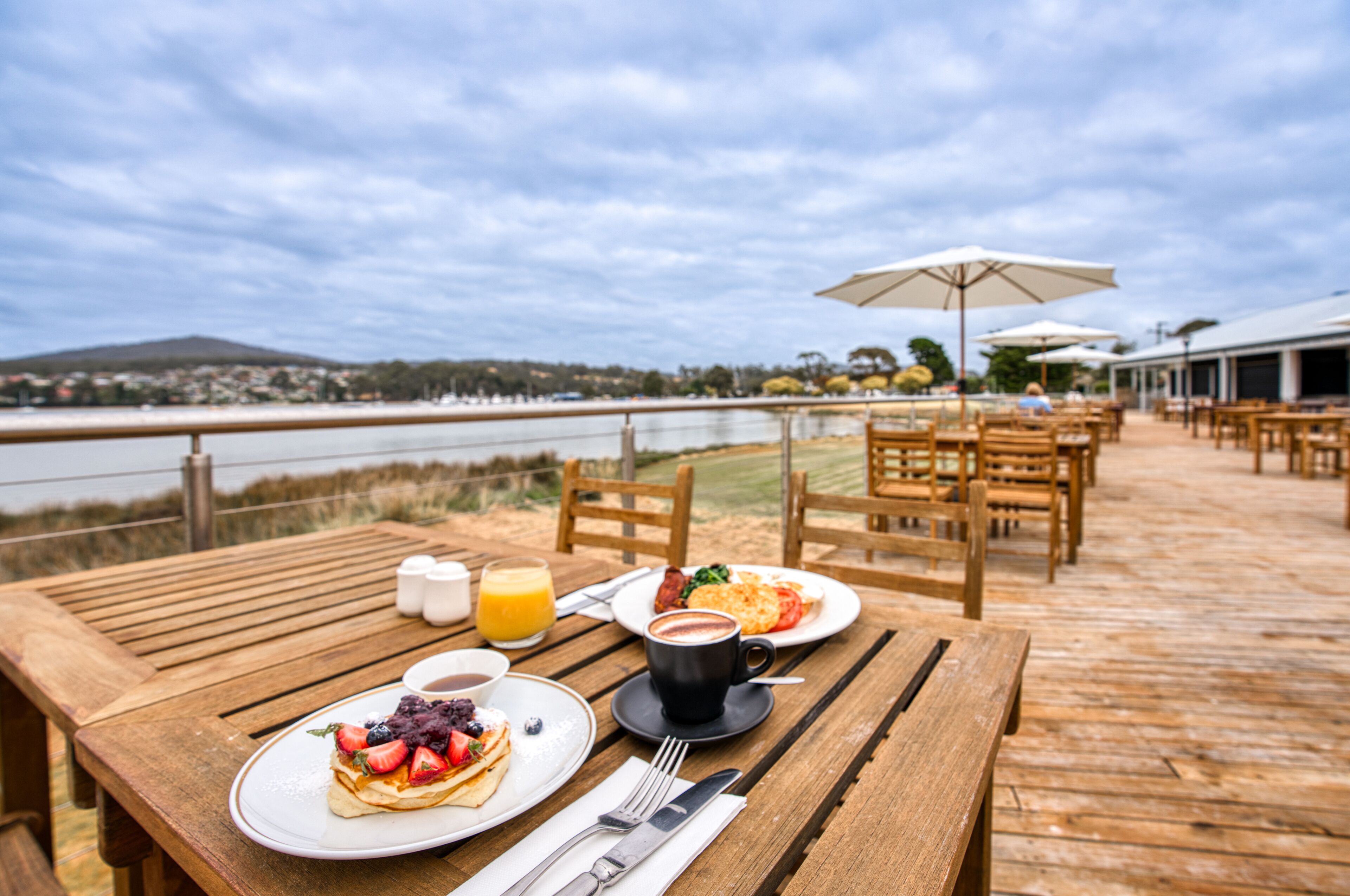 Um deck de madeira com área de estar externa, uma mesa com café da manhã e vista para a água e colinas ao fundo.