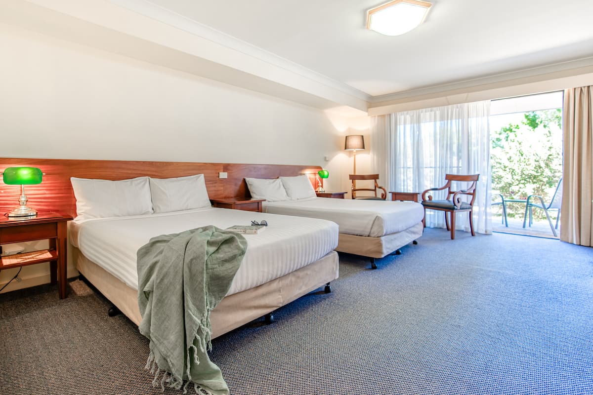 Quarto de hotel com duas camas, um abajur verde, uma cadeira e vista para o verde lá fora.