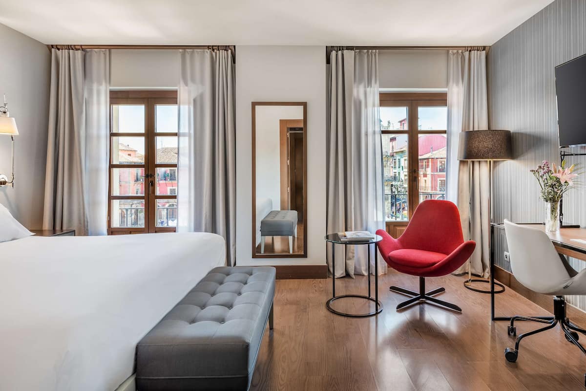 A hotel room with a large bed, a desk with a chair, a television, and a view of the city through the windows.