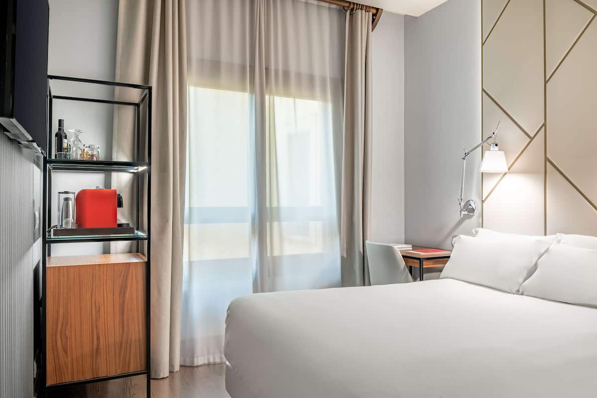 A hotel room with a bed, a nightstand, a lamp, a shelf with toiletries, and a view through sheer curtains.