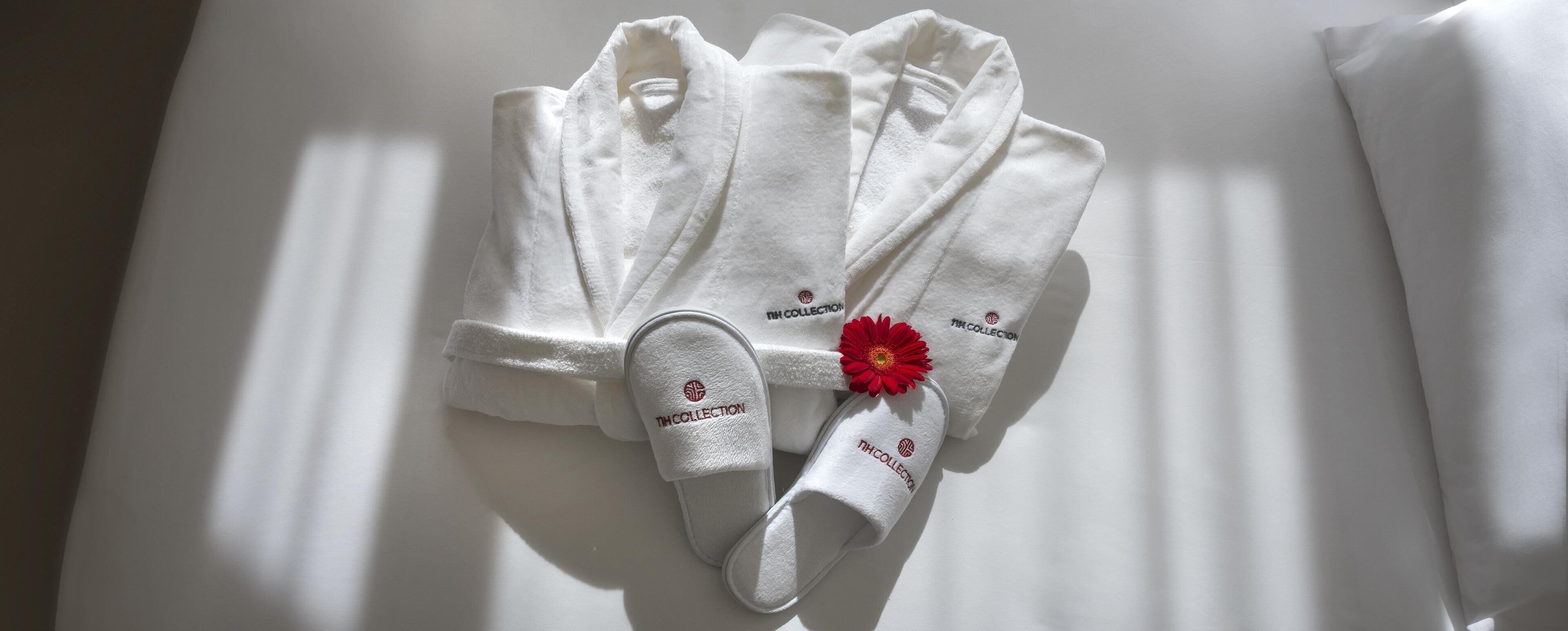Una toalla blanca doblada con cuidado y zapatillas con un adorno de flor roja, con la etiqueta 'NH COLLECTION'.