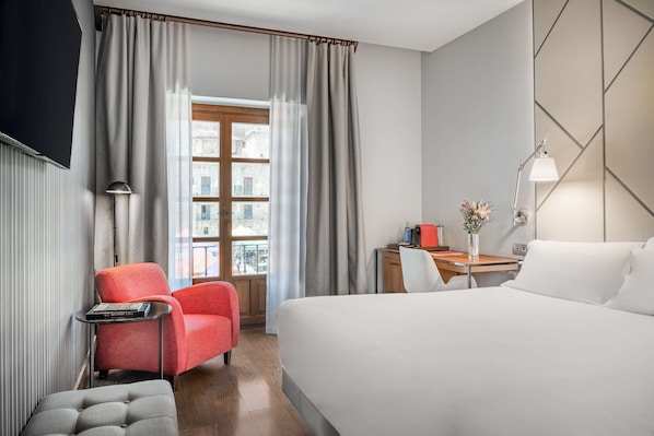 Habitación de hotel con cama, escritorio, silla, lámpara y vista a la ciudad por una ventana.