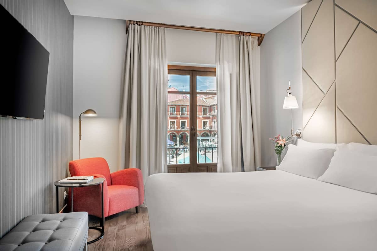 A hotel room with a bed, a red chair, a small table with books, a lamp, and a view of buildings through the window.